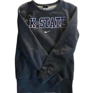 Grå Vintage Sweatshirt - Grå sweatshirt från Nike med K-STATE tryck på bröstet. Tröjan har långa ärmar och en rund halsringning. Perfekt för en avslappnad stil