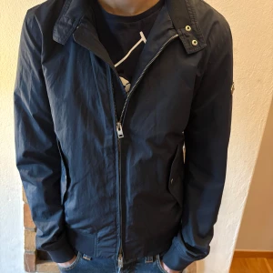 Jacka från J.Lindeberg - Snygg mörkblå jacka från J.Lindeberg med dragkedja och ribbade muddar. Perfekt för en stilren look. Jackan har en diskret logga på ärmen och knappar vid kragen.