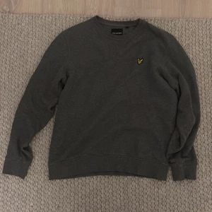 Grå tröja från Lyle & Scott - Säljer en stilren grå tröja från Lyle & Scott med deras ikoniska gula logga på bröstet. Tröjan har långa ärmar och en klassisk rund halsringning. Perfekt för en avslappnad look.
