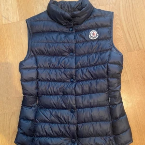 Moncler dunväst  - Säljer en snygg marinblå dunväst från Moncler med knappar framtill. Västen har en quiltad design och en hög krage för extra värme. OBS, liten i storleken!