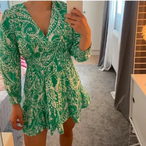 Grön och vit klänning - Säljer en snygg grön och vit klänning med blommigt mönster. Klänningen har en v-ringad design och långa ärmar. Perfekt för vår och sommar med sin luftiga passform och fina detaljer.