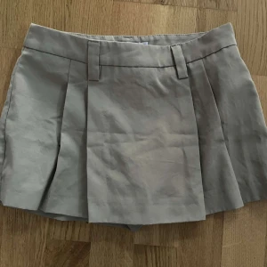 Beige kortkjol med veck  - Snygg beige kortkjol med veck och bälteshällor. Perfekt för en stilren look. Kjolen har en klassisk design som passar till många olika toppar