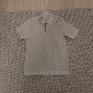 Grå pikétröja från Dobber - Säljer en stilren grå pikétröja från Dobber. Tröjan har en dragkedja framtill och korta ärmar, perfekt för en avslappnad look. Den är tillverkad i ett bekvämt material som passar bra till vardags.