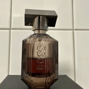 Hugo Boss The Scent Parfym - Elegant parfymflaska från Hugo Boss med en mörk, transparent design. Flaskan har en stilren, geometrisk form och ett metalliskt lock. Doften är en del av 'The Scent'-kollektionen, känd för sina förföriska och maskulina noter. 30 ml