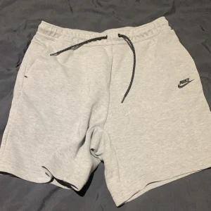 Grå nike tech fleece shorts - Snygga grå shorts från Nike med elastisk midja och dragsko för perfekt passform. De har en diskret Nike-logga på benet och praktiska sidofickor med dragkedja. Perfekta för en avslappnad stil.