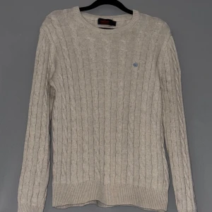 Morris Beige Stickad Tröja - Säljer en stilren beige stickad tröja från Morris med kabelstickat mönster. Tröjan har en rund halsringning och ett diskret broderat emblem på bröstet. Inge skador eller spår av användning finns.