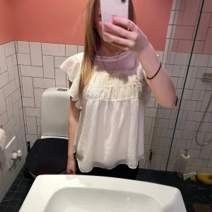 Vit blus med volangdetaljer - Säljer en söt vit blus med volangdetaljer och korta ärmar. Blusen har en luftig passform och är perfekt för varmare dagar. Den har en rund halsringning och fina detaljer.