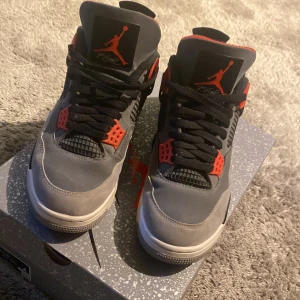 Nike Air Jordan 4 Retro - Snygga Nike Air Jordan 4 Retro i grått med svarta och infraröda detaljer. Skorna har en klassisk design med snörning och den ikoniska Jumpman-loggan på tungan och hälen. Perfekta för sneakerheads som vill ha en stilren look.