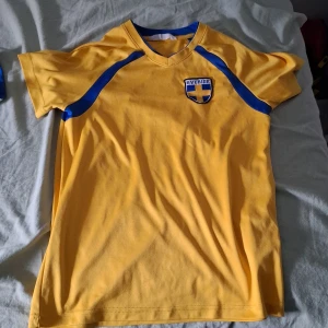 Gul fotbollströja från H&M - Säljer en gul fotbollströja från H&M med blå detaljer och texten 'Sverige' samt nummer 9 på ryggen. Tröjan har korta ärmar och ett emblem på bröstet. Perfekt för fotbollsfans!