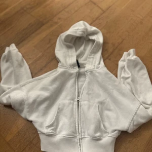 Vit hoodie med dragkedja - Säljer en vit hoodie med dragkedja och fickor framtill. Den har en klassisk design med huva och långa ärmar. Perfekt för en avslappnad stil.