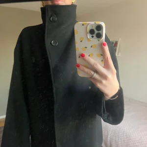 Svart kappa med hög krage - Snygg svart kappa med hög krage och knappar framtill. Där det även går att knäppa upp kragen. Perfekt för kyligare dagar och ger en stilren look. Passar bra till både vardag och mer uppklädda tillfällen.