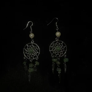 Silvriga drömfångarörhängen med gröna stenar - Handgjorda örhängen i form av drömfångare med silverfärgad metall och gröna kristaller. De har en pärla högst upp och små fjädrar som hänger nedtill. Perfekta för att ge en bohemisk touch till din stil.