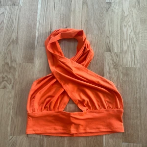 Orange omlott topp - Trendig orange omlott topp med halterneck-design. Toppen är ärmlös och har en öppen rygg vilket ger en snygg och luftig känsla. Perfekt för varma dagar eller en utekväll.