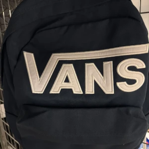 Blå ryggsäck från Vans - Snygg blå ryggsäck från Vans med stort logotyptryck i vitt. Perfekt för att bära dina grejer med stil. Ryggsäcken har ett klassiskt och tidlöst utseende som passar till många tillfällen.