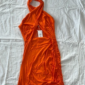 Orange klänning med omlott och rynkade detaljer - Trendig orange klänning med omlott-design och rynkade detaljer på sidan. Den har en unik halterneck-stil och en smickrande passform som framhäver figuren. Perfekt för en somrig look!