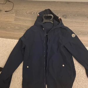 Mörkblå windbreaker från Moncler - Säljer en snygg mörkblå jacka från Moncler med huva och dragkedja. Jackan har en stilren design med Moncler-logga på ärmen och praktiska fickor med knappar. Perfekt för kyligare dagar. Knappt använd och snören medföljer, tog av de pga preferens.