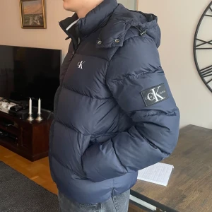 Calvin Klein Pufferjacka - Snygg marinblå pufferjacka i storlek medium från Calvin Klein med dragkedja och avtagbar huva. Perfekt för kyliga dagar.     Använd i ca 4 månader och i gott skick. Om ni vill ha fler bilder eller undrar något mer är det bara att skriva! 🤩
