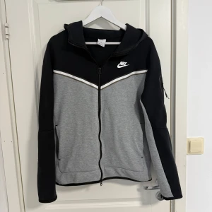 Nike Tech Kofta - Säljer en snygg grå och svart hoodie från Nike med dragkedja och huva. Koftan har en vit Nike-logga på bröstet och en praktisk ficka på ärmen. 