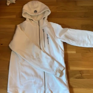 Vit hoodie från Sail Racing - Säljer en stilren vit hoodie från Sail Racing med dragkedja och broderad logga på bröstet. Perfekt för en avslappnad stil. Hoodien har långa ärmar och en bekväm huva.