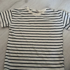 Randig t-shirt från Lindex Kids - Säljer en klassisk vit och svart randig t-shirt från Lindex Kids. Perfekt för barn i åldern 4-6 år. T-shirten har korta ärmar och är tillverkad i ett mjukt och bekvämt material.