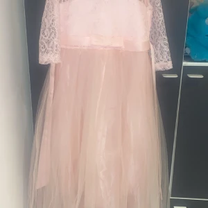 Ljusrosa klänning med spetsdetaljer - Elegant ljusrosa klänning med spetsdetaljer och trekvartsärmar. Klänningen har en vacker tyllkjol och en söt rosett i midjan. Perfekt för speciella tillfällen.