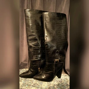 Svarta knähöga stövlar - Sjukt snygga knähöga boots med krokodil-imiterad skinn! De är helt oanvända, kommer tyvärr inte i originalförpackningen, men är felfria och precis som ny. Klackarna är 10 cm höga och bootsen är i wide (för breda fötter).