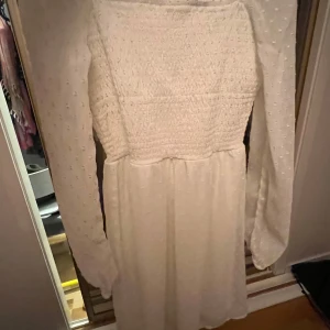 Vit långärmad klänning - Säljer en söt vit klänning med lång ärm och smockdetaljer på överdelen. Klänningen har en luftig passform och är perfekt för vår och sommar. Den har en enkel och stilren design som passar till många tillfällen. BARN STORLEK 11-12 men funkar till vuxna ändå