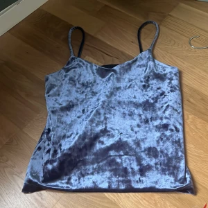 Blå sammetstopp - Säljer en stilren blå sammetstopp med smala axelband. Toppen har en mjuk och glansig yta som ger en lyxig känsla. Perfekt för att lägga till en touch av elegans till din outfit.