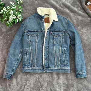 Jeansjacka från Levi's - Säljer en klassisk jeansjacka från Levi's med mjukt teddyfoder. Jackan har en snygg blå tvätt och knappar framtill. Perfekt för kyligare dagar med sin varma insida. Två bröstfickor med knappstängning och en stilren design gör den till ett måste i garderoben.