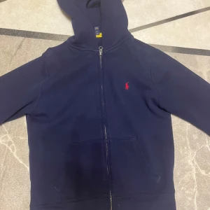 Mörkblå hoodie från Ralph Lauren - Säljer en snygg mörkblå hoodie från Ralph Lauren med dragkedja och huva. Den har en liten röd broderad logga på bröstet och praktiska fickor framtill. Perfekt för en avslappnad stil.