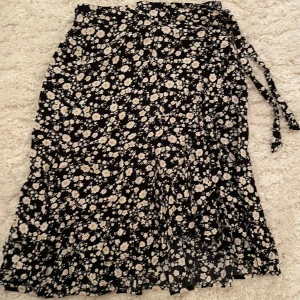 Blommig omlottkjol från Lindex - Säljer en söt blommig omlottkjol från Lindex i svart med vita och gula blommor. Kjolen har ett knytband i midjan och är perfekt för vår och sommar. Den är gjord i ett lätt och bekvämt material. Använd ett fåtal gånger🩷 OBS! Köpare står för frakten!