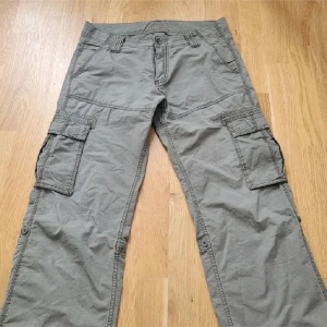 Grå cargopants - Säljer ett par grå/gröna cargopants med flera praktiska fickor och en avslappnad passform. Byxorna har en klassisk design med bälteshällor och knappstängning. 