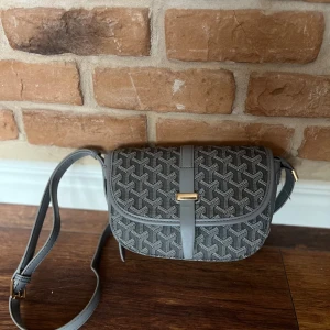 Grå axelväska med mönster - Snygg grå axelväska med ett stilrent geometriskt mönster. Väskan har en justerbar axelrem och en elegant metallspänne framtill. Perfekt för att bära dina nödvändigheter med stil.