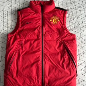 Röd dunväst från Nike med Manchester United-logga - Snygg röd dunväst från Nike med Manchester United-logga på bröstet. Västen har en dragkedja framtill och är perfekt för kyligare dagar. Den har en sportig look och är lätt att matcha med andra plagg. 0 använd gånger.