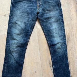 Mörkblå jeans från Nudie Jeans - Snygga mörkblå jeans från Nudie Jeans med klassisk femficksdesign och orange sömmar. De är tillverkade i slitstarkt denim. Perfekta för en avslappnad stil. Storlek 29/34