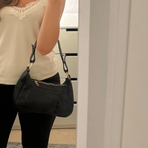 Svart axelväska i syntet - Snygg svart axelväska med krokodilpräglat mönster och gulddetaljer. Väskan har en dragkedja upptill och justerbara remmar. Perfekt för att ge en stilren touch till din outfit.
