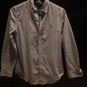 Vit skjorta från Ralph Lauren - Säljer en klassisk vit skjorta från Ralph Lauren med broderad logga på bröstet. Skjortan har långa ärmar och knappar framtill. Perfekt för en stilren look.