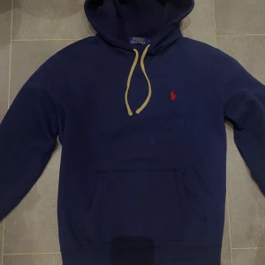 Mörkblå hoodie från Ralph Lauren - Säljer en snygg mörkblå hoodie från Ralph Lauren med det klassiska röda logotypen på bröstet. Tröjan har en känguruficka och justerbar huva med vita snören. Perfekt för en avslappnad stil. Modellen på bilden är 184 cm. Säljs eftersom den passar inte längre. Köptes i december för 2195kr på Zalando nyskick. Snören ser gula ut på bild men kan skicka flera bilder.