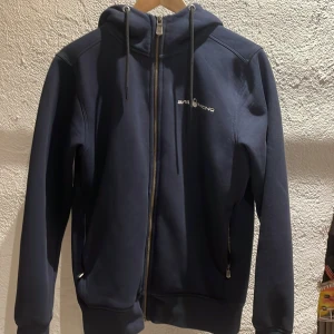 Mörkblå hoodie från Sail Racing - Säljer en mörkblå hoodie från Sail Racing med dragkedja och justerbar huva. Den har en liten logotyp på bröstet och en större på ryggen. Perfekt för en avslappnad stil. 