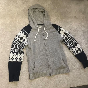 Stickad hoodie med mönster - Säljer en riktigt fet stickad grå hoodie  med mörkblåa och vita mönstrade armar. Perfekt till vinter och vår!