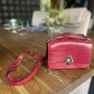Röd axelväska i krokodilmönster - Snygg röd axelväska med krokodilmönster. Väskan har en justerbar axelrem och ett handtag för flexibel användning.