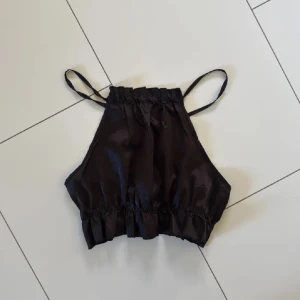 Satin topp - Snygg svart satin topp med smala axelband och volangdetalj vid nederkanten. Perfekt för en stilren look. Justerbara axelband för bästa passform.