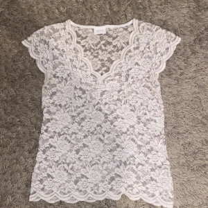 Vit spetsblus från Vila - Säljer en elegant vit spetsblus från Vila med vackert blommönster. Blusen har korta ärmar och en v-ringad design med fina vågiga kanter. Perfekt för en stilren look.