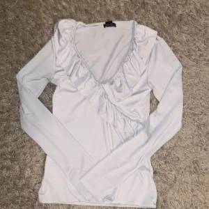 Vit långärmad topp från Amisu - Säljer en stilren vit långärmad topp från Amisu. Toppen har en v-ringad design med volangdetaljer längs halsringningen och framsidan. Perfekt för en elegant look.