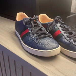 Gucci skor - Snygga blå sneakers med präglat mönster och klassiska röd-gröna ränder på sidan. Skorna har en unik röd detalj på hälen och snörning framtill. Perfekta för en stilren look.