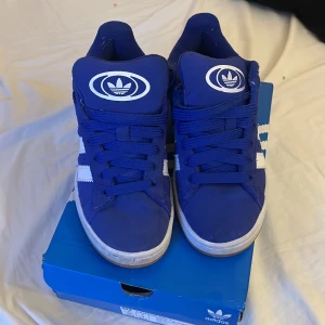 Blå Adidas Campus sneakers - Säljer ett par blå Adidas Campus sneakers med vita detaljer och klassiska tre ränder på sidan. Skorna har snörning och en bekväm Ortholite-innersula. Perfekta för en sportig och stilren look.
