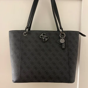 Svart axelväska från Guess - Snygg svart axelväska från Guess med subtilt mönster och metallogo framtill. Väskan har dubbla handtag och en dragkedja baktill för extra förvaring. Insidan är rymlig med flera fack och Guess-logga på fodret. Perfekt för att bära med sig allt man behöver. Nypris är 3000 kr. 
