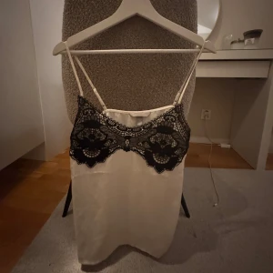 Vit linne med svart spetsdetalj - Elegant vit linne med tunna axelband och vacker svart spetsdetalj över bysten. Perfekt för vardag eller fest!