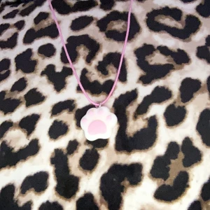 Rosa halsband med tassberlock - Sött halsband med en rosa tassformad berlock. Berlocken hänger på ett rosa snöre och ger en lekfull touch till din stil. Perfekt för djurälskare eller som en rolig accessoar.