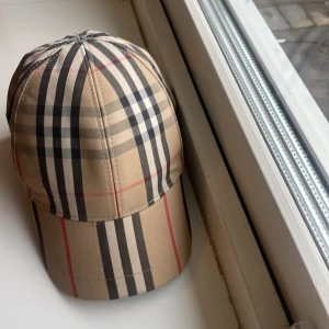 Rutig keps från Burberry - Snygg keps från Burberry i klassiskt rutigt mönster med beige, svart och röd färg. Kepsen har justerbar passform och är perfekt för en stilren look. Kepsen är till salu för att den blivit för liten och är självklart äkta. Köptes på NK och är i storlek 54cm